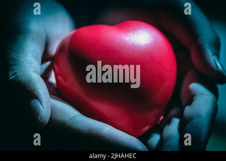 Gros plan des mains tenant le cœur rouge ou la forme de l'amour dans les paumes sur un arrière-plan sombre. Concept de condoléances et de sympathie. Banque D'Images