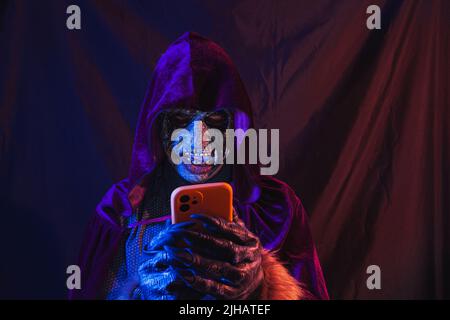 Portrait d'un zombie habillé dans une chemise et cape à capuchon à l'aide de son téléphone cellulaire. La scène est sombre, éclairée par des lumières bleues et orange, le visage du monstre est Banque D'Images