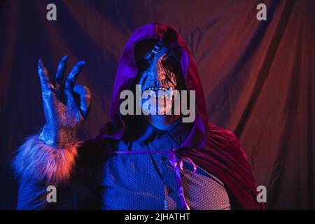 Portrait d'un zombie habillé dans une chemise et cape à capuche faisant un geste droit avec sa main. La scène est sombre, éclairée par des lumières bleues et orange Banque D'Images