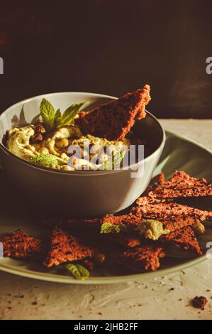 Houmous d'avocat dans un bol avec pois chiches, feuilles de menthe et craquelins faits maison sur fond blanc. Aliments végétaliens crus sains Banque D'Images