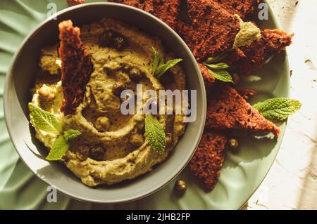 Houmous d'avocat dans un bol avec pois chiches, feuilles de menthe et craquelins faits maison sur fond blanc. Aliments végétaliens crus sains Banque D'Images