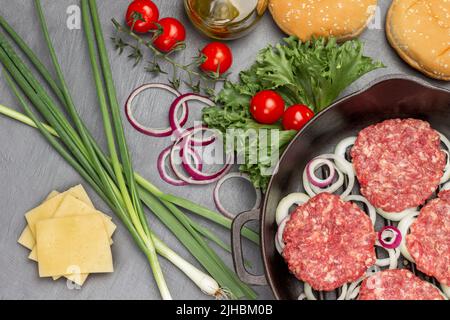 Dans une poêle, faire cuire des hamburgers crus avec des oignons. Fromage, oignons verts, brins de tomate et laitue sur la table. Pose à plat. Arrière-plan gris Banque D'Images