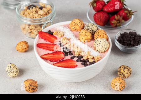 Bol à smoothie avec fraises, granola, chocolat, flocons de noix de coco et boules de miel croquantes dans un bol blanc sur fond gris Banque D'Images