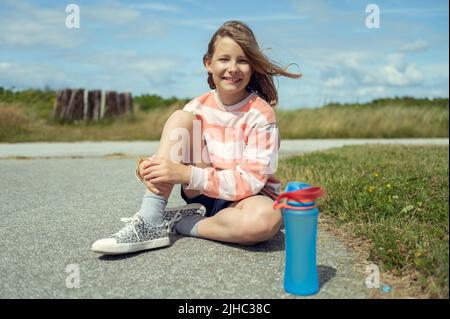 Portrait d'une jeune fille adorable avec un beau sourire assis sur le sol dans des baskets avec une bouteille d'eau bleue pendant les vacances d'été Banque D'Images