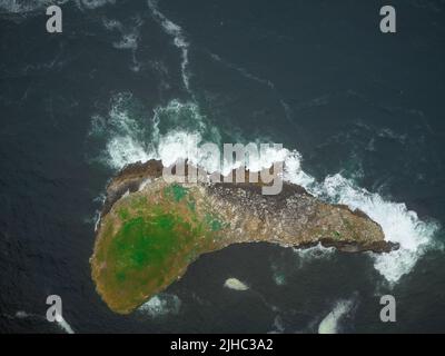 Vue aérienne. Une île en pierre dans l'océan qui ressemble à un gros poisson. Dans les endroits surcultivés avec de la mousse verte. Eau turquoise foncé et vagues de mousse blanches. Banque D'Images