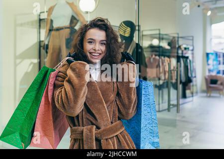 Soldes des fêtes. Belle jeune femme en fourrure manteau tenant des sacs de papier de couleur avec des achats et des cadeaux pour les vacances de Noël pour la famille et les amis. Spectacles, brags, réjouissances, regarde la caméra dans un magasin dans un centre commercial Banque D'Images