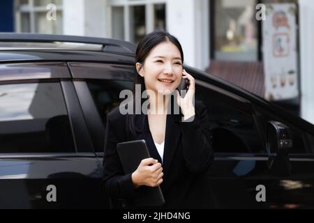 Les hommes d'affaires asiatiques, les propriétaires d'entreprise, les présidents d'entreprise ou les femmes d'employés qui parlent au téléphone et tiennent une tablette sortent de leur voiture Banque D'Images