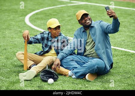 Papa heureux faisant le portrait de selfie avec son fils sur son téléphone mobile pendant qu'ils reposent sur l'herbe après le match de baseball Banque D'Images