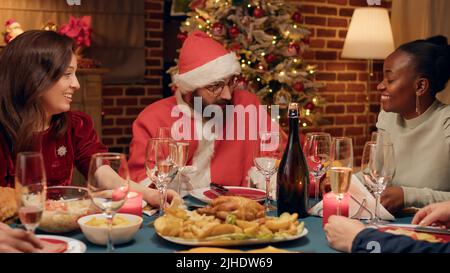 Drôle de mari déguisé comme le Père Noël discutant avec les gens tout en célébrant les vacances d'hiver. Homme festif portant le costume du Père Noël lors d'un dîner de Noël en discutant avec des membres de la famille. Banque D'Images