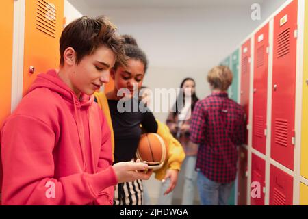 Les jeunes élèves du secondaire se tenant près d'un casier dans le couloir du campus qui parlent et utilisent un téléphone intelligent. Banque D'Images