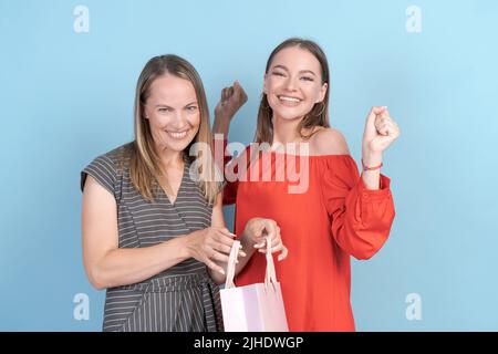 Mère et fille tiennent des sacs d'achats et des cadeaux. Joyeuse fille sourit joyeusement de ce qu'elle voit dans le paquet, dans une robe grise et rouge sur un fond bleu. Se gestuelle émotionnellement Banque D'Images