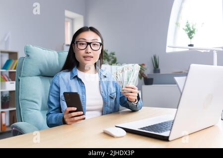 Formation professionnelle sur l'argent. Une jeune femme d'affaires asiatique organise une session de formation sur le thème de l'argent. Il tient un téléphone et une liade de factures. Assis à son bureau, parle, regarde dans la caméra. Banque D'Images