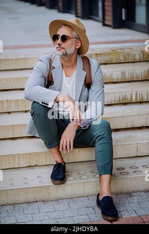 Homme confiant portant un chapeau de paille et un sac à dos assis dans les escaliers, homme d'affaires dans des vêtements décontractés en été sur le chemin du travail. Banque D'Images