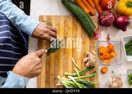 Section médiane à angle élevé de la femme biraciale mature portant un tablier coupant des légumes à bord dans la cuisine Banque D'Images