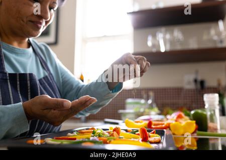 Section médiane de la femme biraciale mûre portant un tablier parsemant les épices sur les légumes dans le plateau de la cuisine Banque D'Images