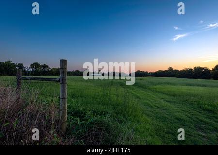 Paysage de campagne de voies de roue à travers un champ de foin vert menant à une grange de pôle au loin au coucher du soleil. Banque D'Images