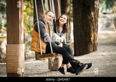 Vue latérale d'un jeune couple mignon en balançoire en automne dans des vêtements chauds aux couleurs de base dans le parc à l'extérieur Banque D'Images