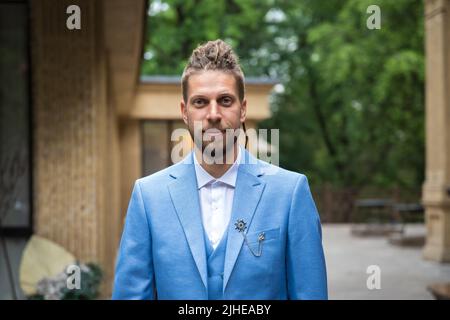 Portrait d'un jeune homme de 25 ans avec une coiffure hippster et une barbe en costume bleu trois pièces dans un espace de restaurant dans un parc Banque D'Images