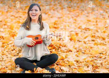 Jeune femme asiatique joyeuse jouant ukulele dans le parc d'automne sur les feuilles mortes Banque D'Images