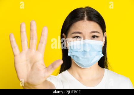 Demander d'arrêter la femme asiatique d'âge moyen portant un masque médical gesturant pour rester à l'extérieur avec des yeux positifs sur l'appareil photo isolé sur fond jaune. Le concept de pandémie, la varicelle ou le coronavirus. Banque D'Images