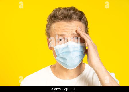 Le jeune homme souffrant d'un mal de tête tient sa tête portant un masque médical sur son visage. Un jeune homme malade en masque facial ressent une migraine touchant sa tête. Un homme caucasien malade en masque de protection. Banque D'Images