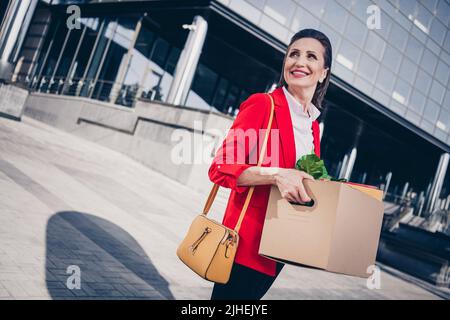 Photo de brillant bonne humeur mature femme enseignant porter une veste rouge tenant des affaires laissant le travail à l'extérieur de la ville complexe d'affaires Banque D'Images