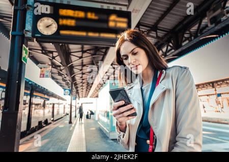 Une jeune femme avec des heures de départ derrière elle attendant son train tout en tenant son téléphone mobile - une femme regardant l'horloge dans la gare Banque D'Images