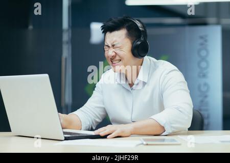 Homme d'affaires asiatique regardant un match de sport au bureau, homme regardant déçu par l'ordinateur portable, utilisant des écouteurs pour écouter Banque D'Images