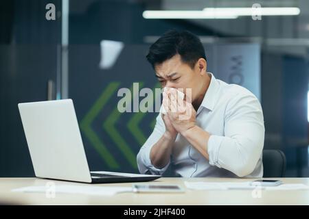 Malade dans le bureau tousse et a le nez qui coule, homme d'affaires asiatique avec froid travaille dans le bureau sur ordinateur portable Banque D'Images