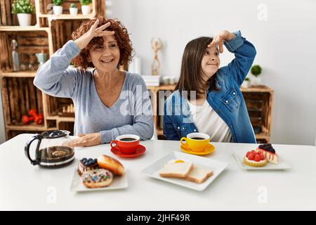 Famille de la mère et fille de syndrome de Down assis à la maison manger le petit déjeuner très heureux et souriant regardant loin avec la main sur la tête. Recherche co Banque D'Images