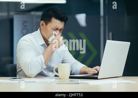 Malade dans le bureau tousse et a le nez qui coule, homme d'affaires asiatique avec froid travaille dans le bureau sur ordinateur portable Banque D'Images