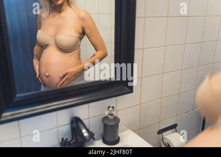 Personne enceinte caucasienne non reconnaissable debout devant le miroir dans la salle de bains, regardant le ventre de la femme enceinte tout en portant un soutien-gorge sans bretelles. Photo de haute qualité Banque D'Images