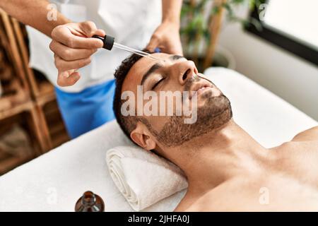 Deux hommes hispaniques thérapeute et patient ayant un traitement du visage en utilisant le sérum au centre de beauté Banque D'Images