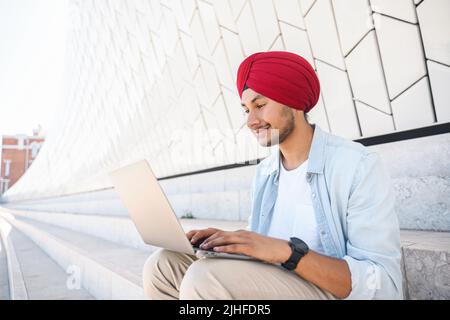 Vue latérale d'un jeune travailleur indépendant indien moderne dans le turban national utilisant un ordinateur portable, un homme de développement hindou dans une chemise décontractée dactylographiant assis à l'extérieur dans le paysage urbain, dorant, travaillant à distance sur l'externalisation Banque D'Images