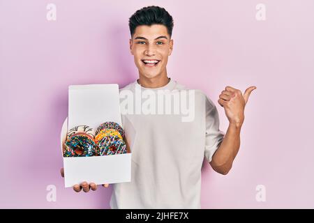 Jeune homme hispanique tenant une boîte de beignets colorés savoureux pointant le pouce vers le côté souriant heureux avec la bouche ouverte Banque D'Images