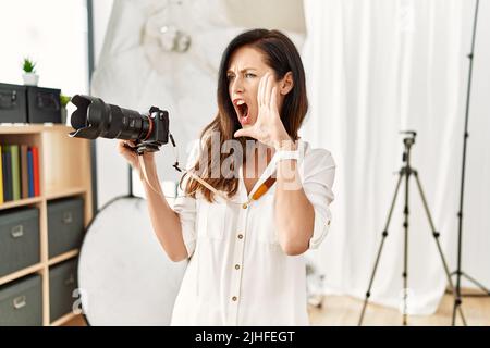 Belle femme caucasienne travaillant comme photographe au studio de photographie criant et criant à voix haute à côté de la main sur la bouche. Concept de communication. Banque D'Images