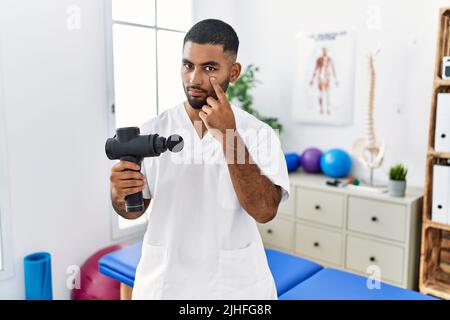 Jeune kinésithérapeute indien tenant un pistolet de massage thérapeutique au centre de bien-être pointant vers l'œil en regardant vous geste, expression suspecte Banque D'Images
