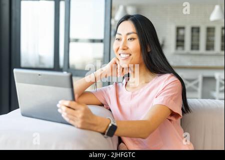 Femme asiatique utilisant un coussin moderne tout en étant assise à la maison, souriant, parlant par vidéo, se détendre sur un grand canapé gris tout en surfant sur le Web sur une tablette numérique, faire une photo de sefie. Surfez sur le Web. Banque D'Images