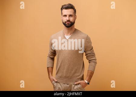 Beau calme brunette homme portant des vêtements décontractés regardant l'appareil photo en gardant les mains dans les poches isolées sur fond beige Banque D'Images