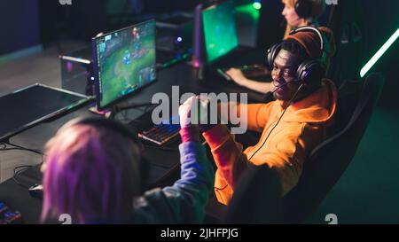 Un homme à la fois décontracté et barbu, vêtu d'un casque, jouant à un jeu d'équipe en ligne populaire et faisant une bosse de poing avec un autre joueur. Photo de haute qualité Banque D'Images