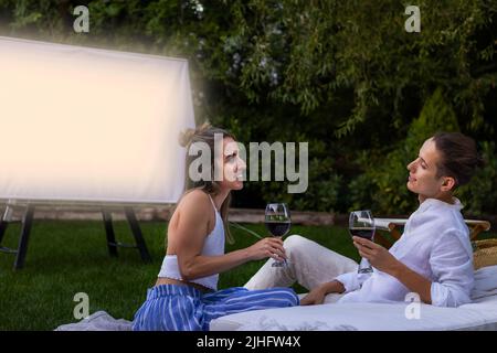 Jeune mignon en couple amoureux ayant une date dans le jardin avec un verre de vin tout en regardant un film à l'extérieur en été Banque D'Images