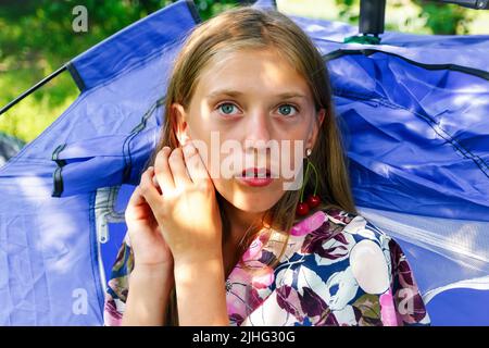 Refocalisation belle blonde jeune fille avec cerise douce sur l'oreille. Belle adolescente souriante à proximité, contre le vert du parc d'été. Tente de camping. Sur Banque D'Images