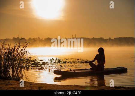 Silhouette d'une femme en position lotus sur un panneau gonflable SUP à l'aube et à l'horizon avec des brouillards sur l'eau Banque D'Images
