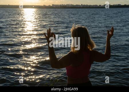 Silhouette d'une femme tenant ses doigts dans une mudra pose sur le fond de l'eau de la mer, lac. Méditation. Sports dans la vieillesse. Yoga Banque D'Images