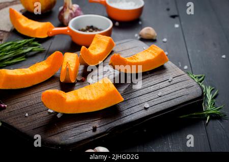 morceaux de citrouille crus sur une planche à découper avec romarin, sel, épices sur une table sombre ancienne. Légumes préparés pour cuire au four. Automne cuisine maison Banque D'Images