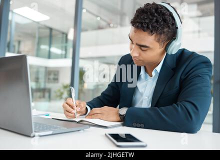 Jeune homme d'affaires sérieux de course mixte portant des écouteurs à l'écoute de la musique tout en écrivant des idées dans un seul ordinateur portable au travail. Un mâle hispanique Banque D'Images