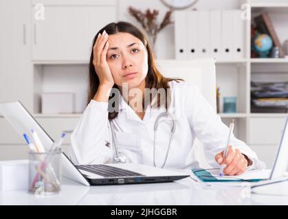 Inquiète femme médecin travaillant à l'ordinateur portable avec des papiers dans le bureau Banque D'Images