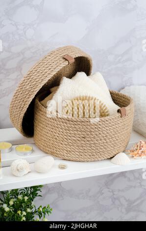 Panier en jute tricoté à la main avec un couvercle avec des serviettes et une brosse dans la salle de bains sur un rack en bois blanc. Banque D'Images