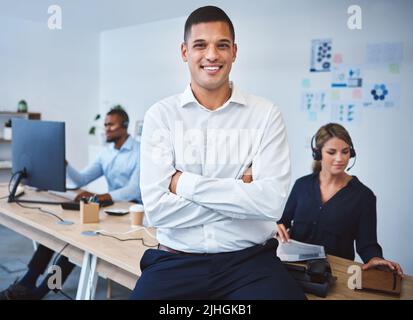 Portrait d'un jeune agent de télémarketing de centre d'appels de course mixte confiant debout avec les armes croisées tout en travaillant dans un centre d'appels avec des collègues dans Banque D'Images