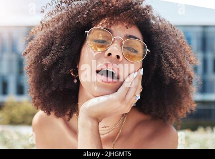 Portrait d'une jeune femme branchée de race mixte avec un afro souriant et posant seul à l'extérieur. Femme hispanique portant des lunettes de soleil et se sentant heureuse Banque D'Images